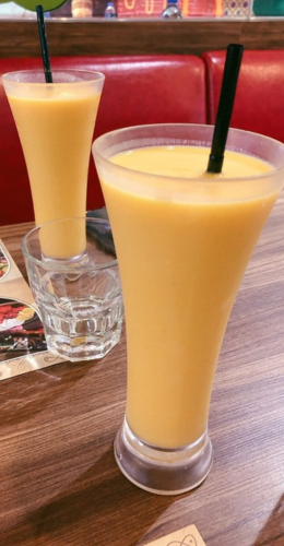 The are two straw-equipped drinks on an eating surface with nibbles, drinks, including juices