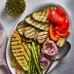 A bowl on a wooden table with a spoon resting over a plate of vibrant and varied veggies